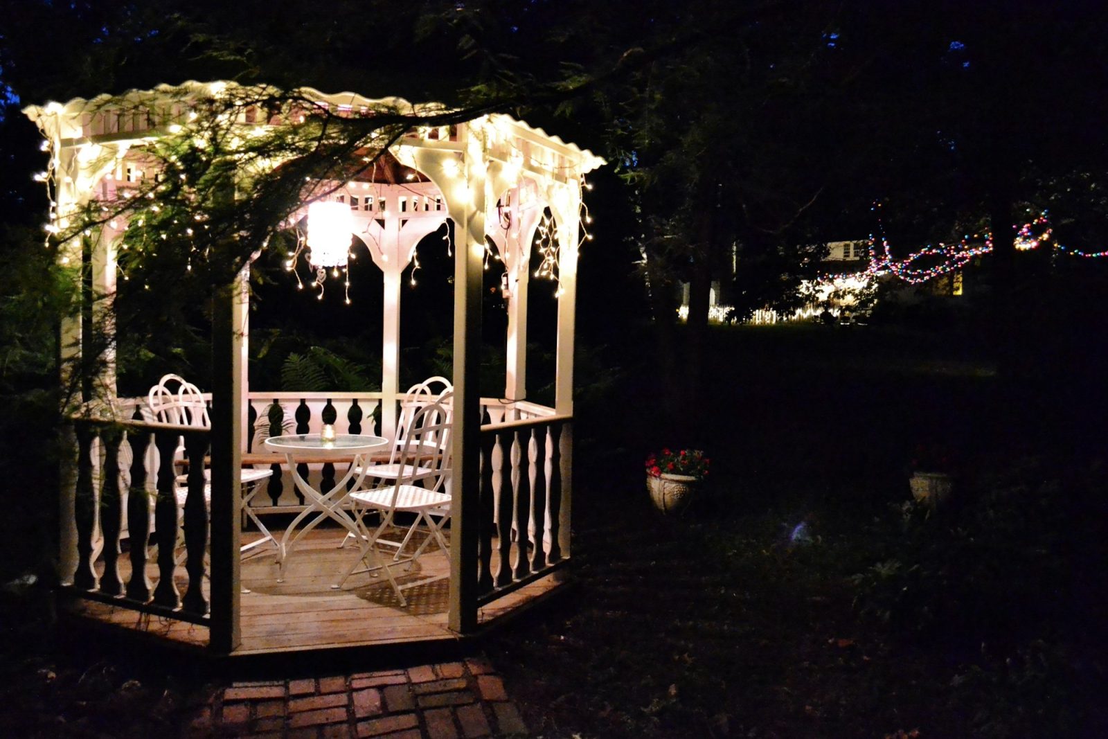 Gazebo in a garden at night with lights, a table and chairs with house party lights in background.